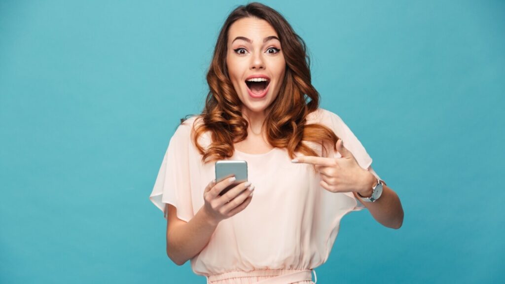 Retrato de uma mulher animada usando um vestido, apontando o dedo para o telefone, isolada sobre um fundo azul.