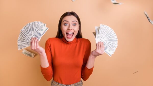 mulher com expressão facial animada segurando dois le de dinheiro, um em cada mão, em fundo pêssego