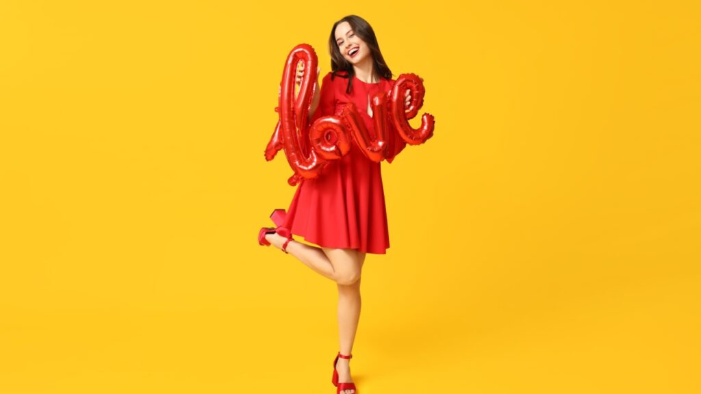 no centro da imagem há uma mulher com vestido vermelho e segurando um balão vermelho que forma a palavra love em fundo amarelo