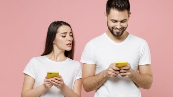 à esquerda está uma mulher segurando um celular mas com a cabeça empinada para a direita olhando o celular do homem que está com lado dela. Os dois usam branco e estão em fundo rosa