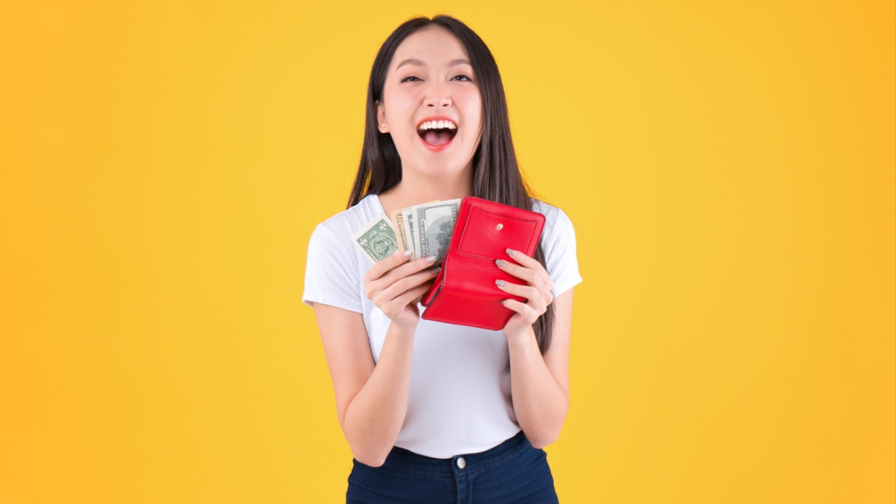mulher com traços asiáticos segurando uma carteira vermelha e tirando dinheiro dela em fundo amarelo