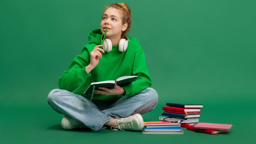 Retrato de uma jovem estudante sentada no chão uma com expressão pensativa, estudando, isolada sobre um fundo verde de um estúdio. Conceito de educação, estudo, lição de casa, juventude, estilo de vida.