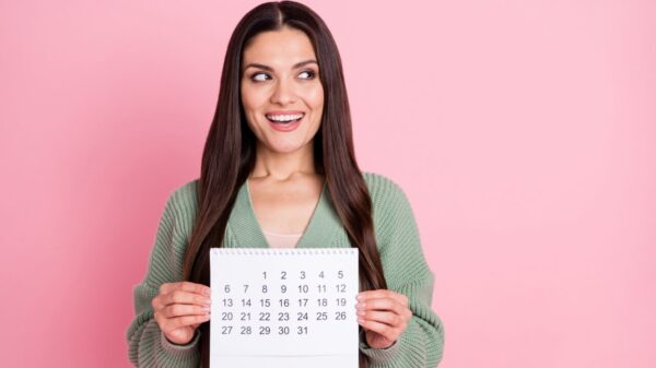 Foto de uma mulher mostrando um calendário, olhando para o lado, usando uma roupa verde, isolada em um fundo de cor rosa pastel.