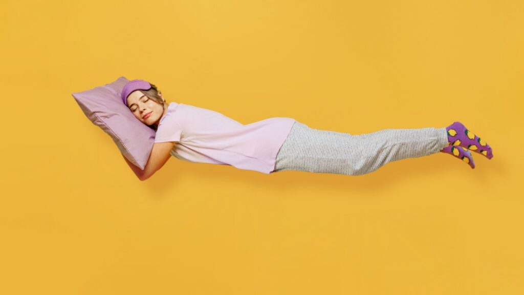 mulher vestindo pijama em tons de roxo. ela está deitada com a cabeça apoiada em um travesseiro roxo e o fundo da imagem é amarelo
