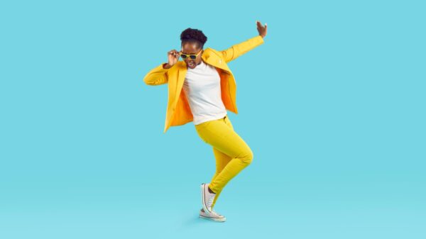 Ao centro uma mulher negra e usando usando um conjunto amarelo em posição de dança em fundo azul