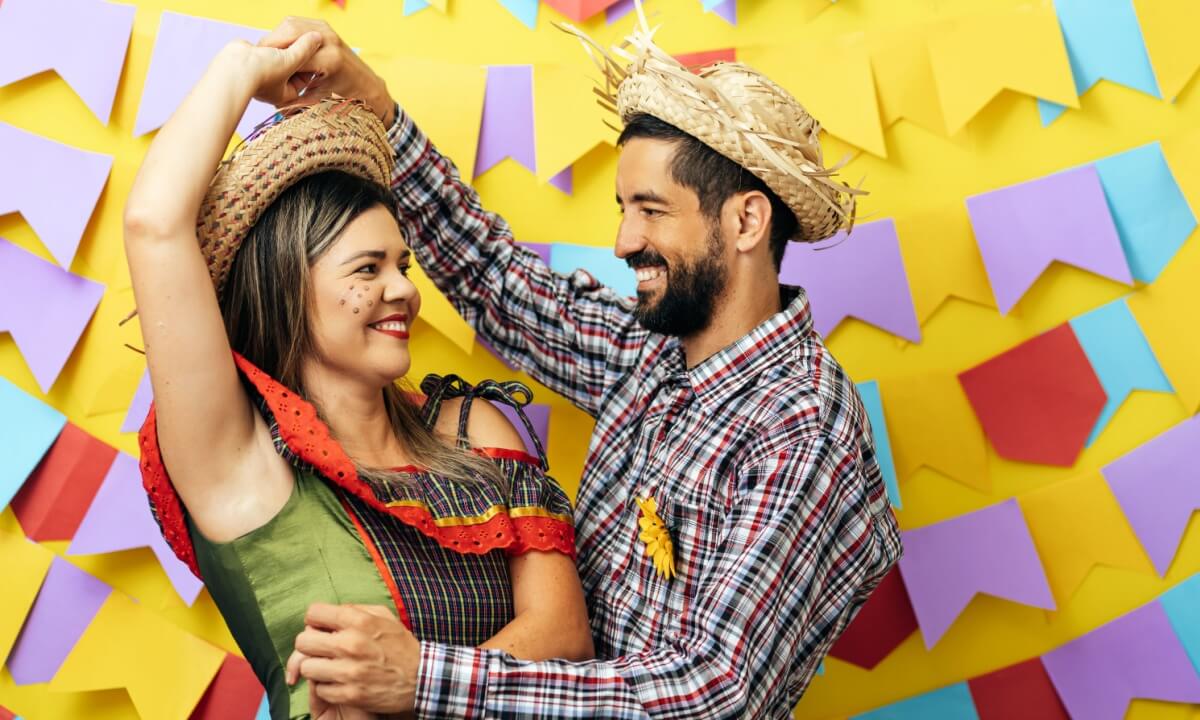 casal fantasiado com roupas de festa junina na frente de um fundo colorido com bandeirinhas juninas