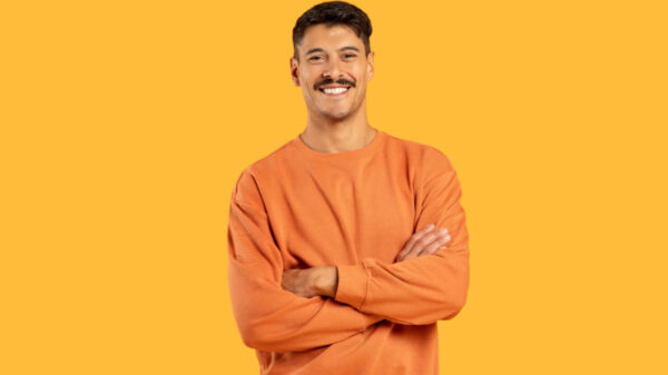 Homem alegre com os braços cruzados, confiante, usando um moletom laranja casual, contra um fundo amarelo.
