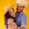 Casal vestindo roupas tradicionais de festa junina dançando em um fundo amarelo.