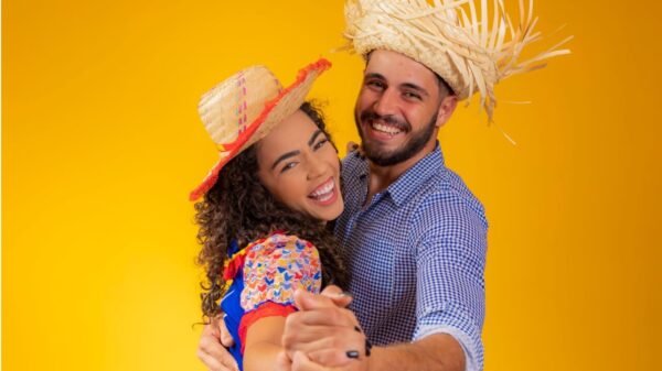 Casal vestindo roupas tradicionais de festa junina dançando em um fundo amarelo.