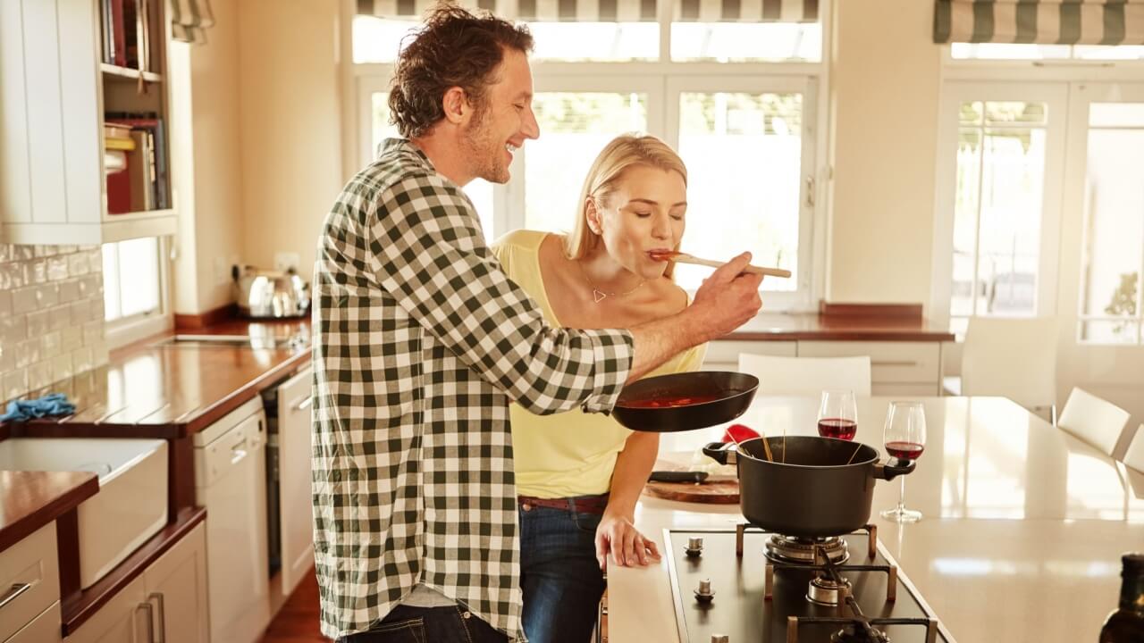 casal cozinhando na cozinha onde ele está segurando uma colher de madeira e colocando na boca da mulher para ela experimentar