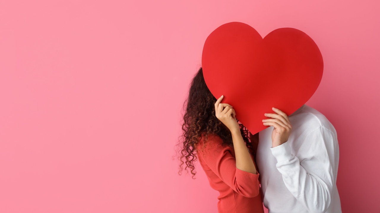 casal no canto direito com imagem se beijando com os rostos escandidos com um grande coração vermelho de cartolina em fundo rosa