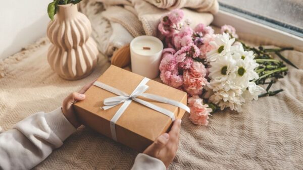 mão feminina segurando uma caixa alaranjada de presente com um laço e flores rosa ao lado