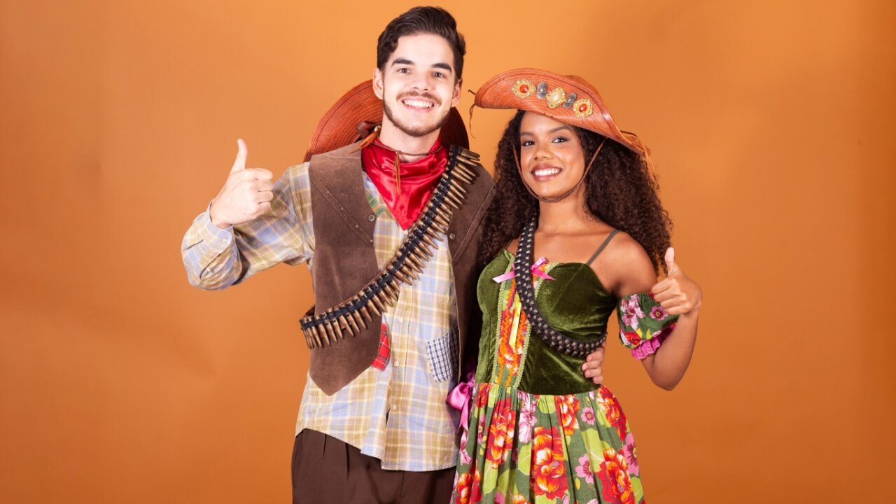 casal com roupas típicas de festa junina abraçados e fazendo o sinal de joia com a mão em fundo laranja