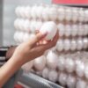 mão feminina segurando um ovo branco com caixas transparentes de ovos ao fundo