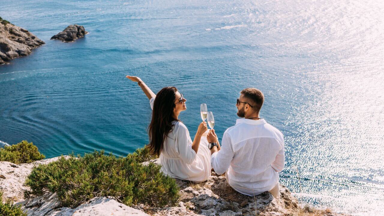 fotografia tirada de cima de um casal sentado em uma pedra brindando champanhe e o mar ao fundo