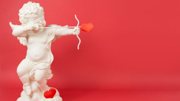à esquerda está uma estátua branca do cupido atirando uma flecha em forma de coração em fundo todo vermelho