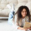 mulher sorrindo lendo um livro deitada no tapete com um sofá atrás