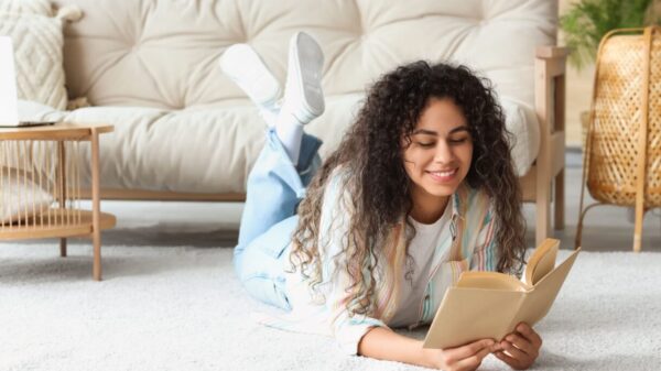 mulher sorrindo lendo um livro deitada no tapete com um sofá atrás