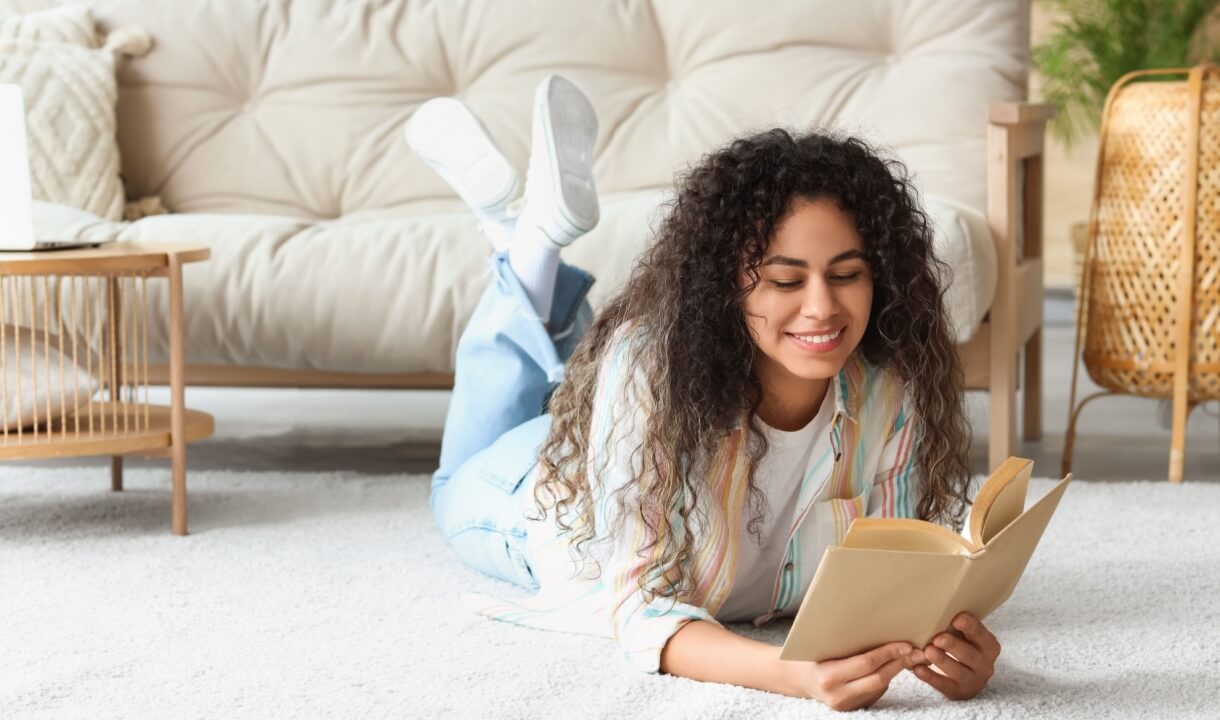 mulher sorrindo lendo um livro deitada no tapete com um sofá atrás