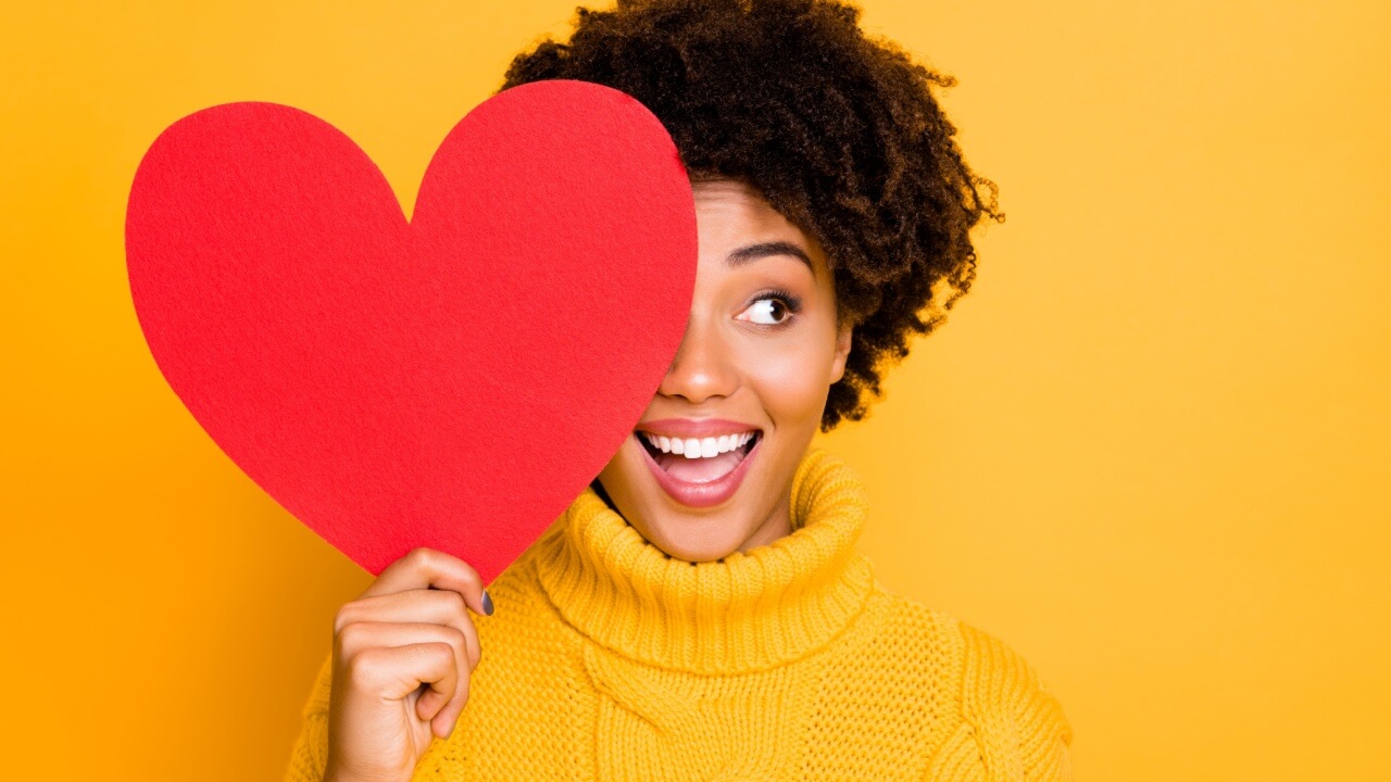 mulher negra vestindo roupa de frio amarela em fundo amarelo segurando um coração vermelho grande na altura do olho