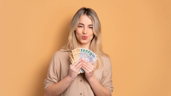 mulher segurando um leque com notas de cem e cinquenta reais em fundo pêssego