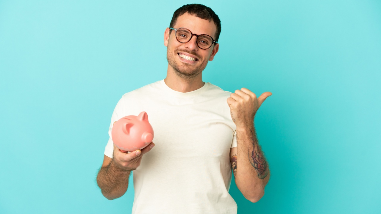 homem jovem com camisa branca segurando um porquinho de cofre em fundo azul claro