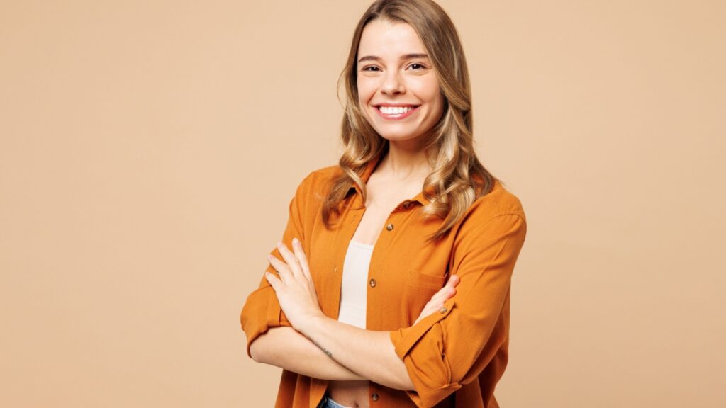 no centro da imagem está uma mulher jovem sorrindo de braços cruzados em fundo marrom alaranjado