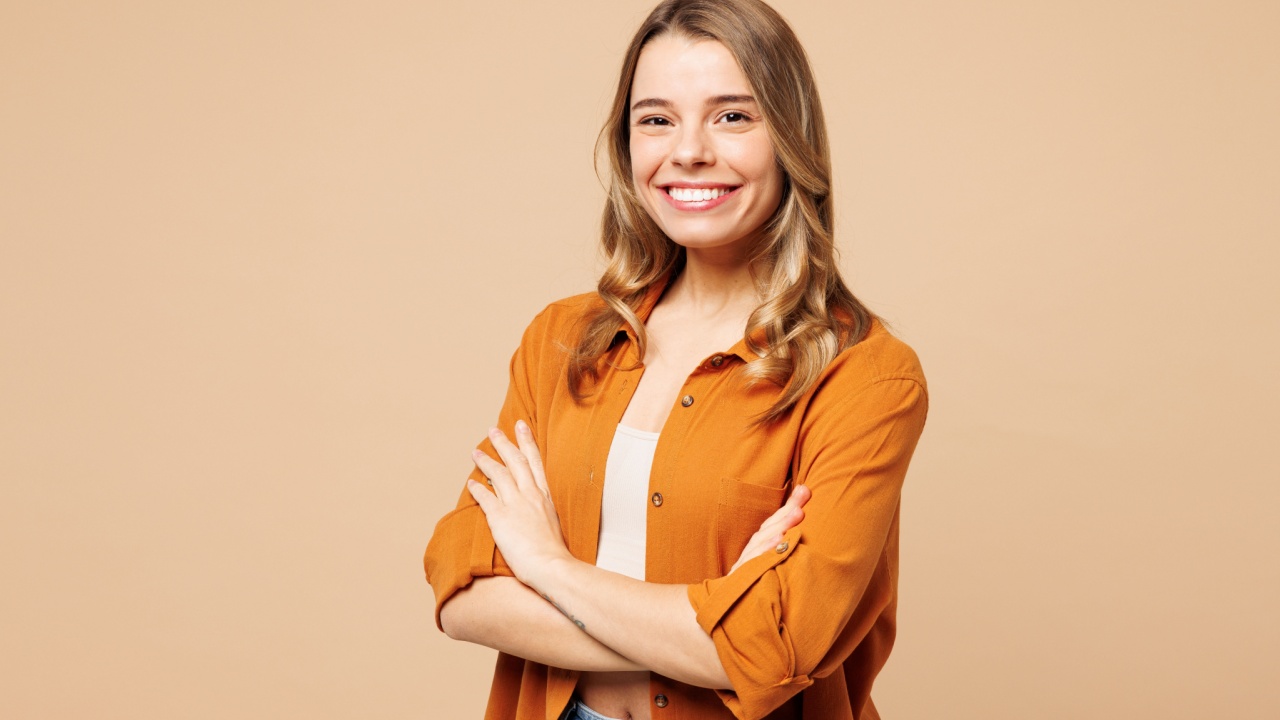 no centro da imagem está uma mulher jovem sorrindo de braços cruzados em fundo marrom alaranjado