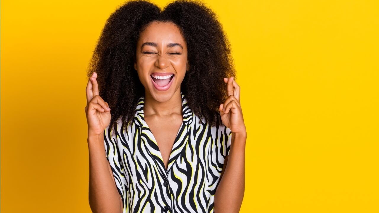 Foto de mulher de cabelos encaracolados usando uma camisa com estampa de zebra, com os dedos cruzados, torcendo por sorte, isolada em um fundo amarelo.