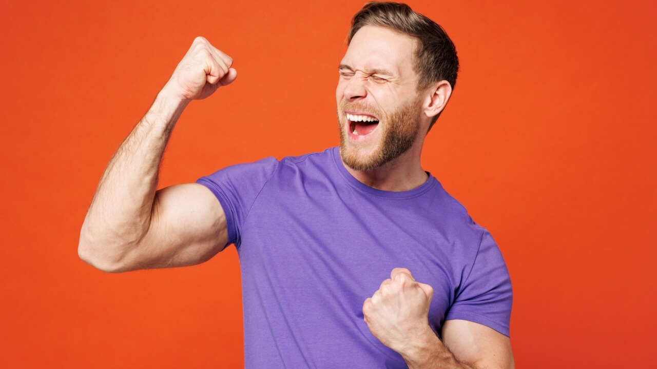 Homem animado, muito feliz, usando uma camiseta casual roxa, fazendo gesto de vencedor, comemorando, com os punhos cerrados, isolado em um estúdio com o fundo de parede laranja. Conceito de estilo de vida.