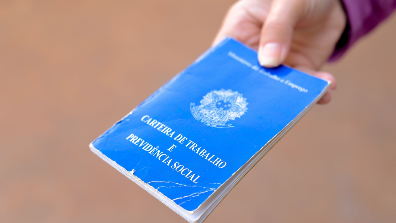 mão masculina branca segurando uma carteira de trabalho em fundo marrom