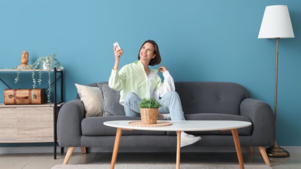Mulher sorrindo, segurando um controle remoto de ar condicionado, sentada em um sofá encostado em uma parede azul.