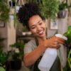 mulher negra sorrindo e regando planta numa sala cheia de plantas e flores