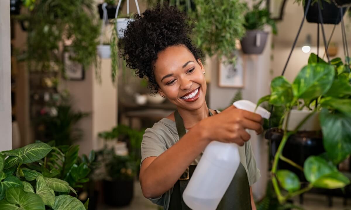 mulher negra sorrindo e regando planta numa sala cheia de plantas e flores