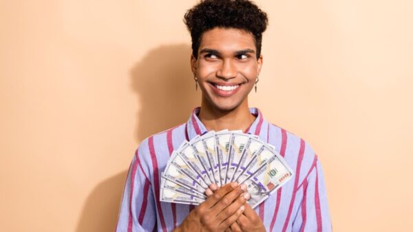 Foto de um rapaz jovem em um espaço vazio segurando notas de dinheiro, usando uma roupa elegante e listrada, isolado em um fundo bege.