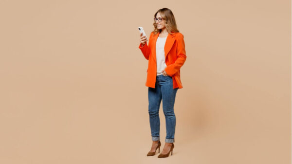 Corpo inteiro de uma mulher feliz usando ma roupa formal com um terno laranja, usando um óculos, segurando na mão um celular, isolada em estúdio com um fundo bege simples.