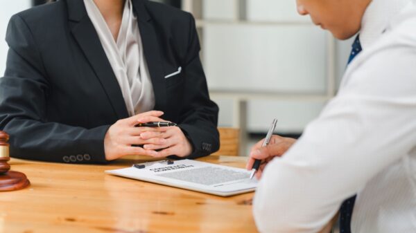 duas pessoas sentadas em uma mesa de madeira e um deles está assinando um contrato