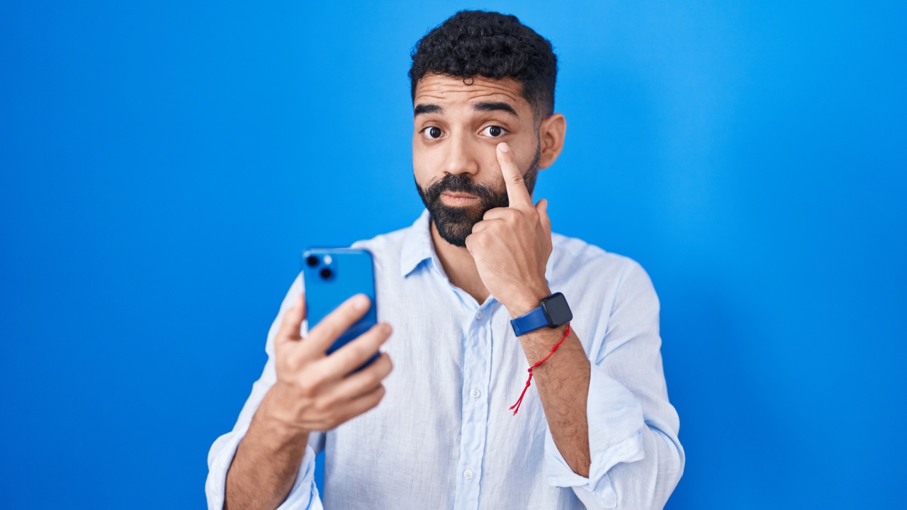 homem com o dedo indicador apontado para o olho em fundo azul escuro e segurando um iphone da mesma cor