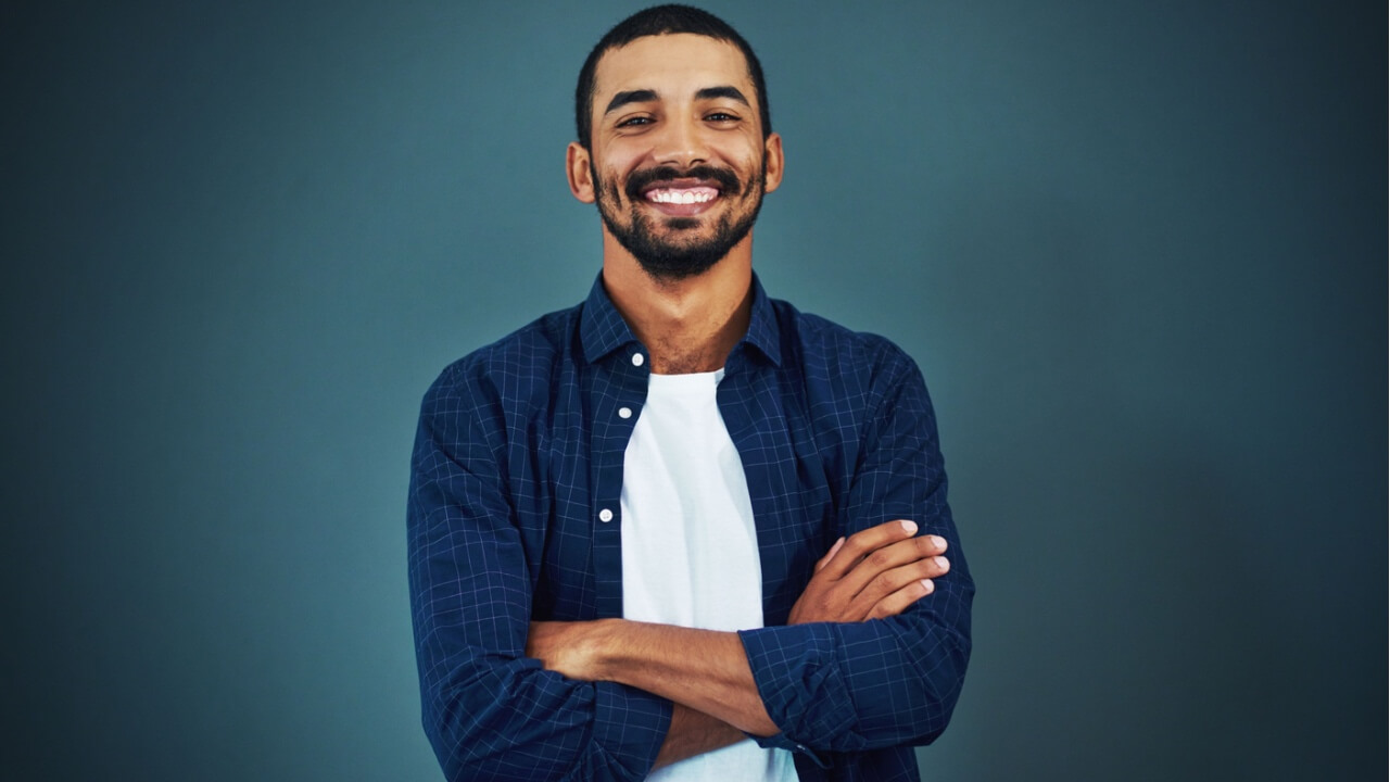 Retrato de um homem de braços cruzados, feliz, sorrindo e confiante em um estúdio, isolado em um fundo escuro.