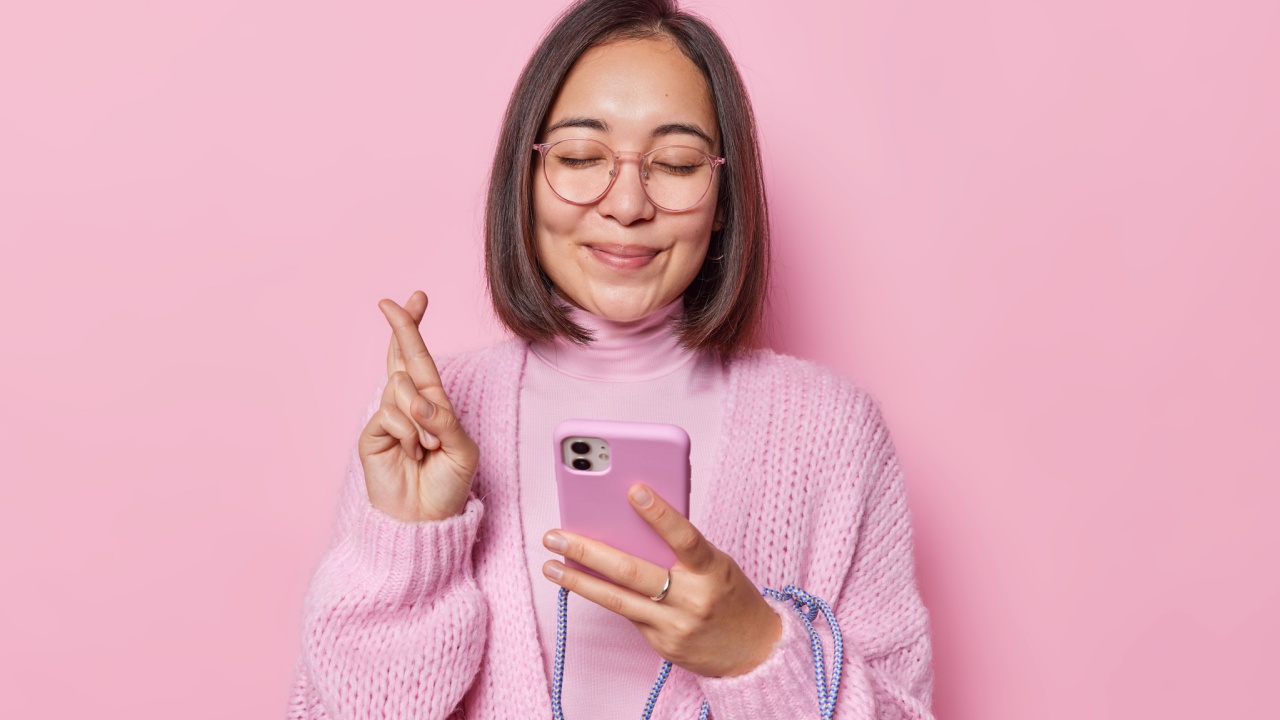 mulher com traços asiáticos vestindo rosa com os dedos cruzados em fundo também rosa