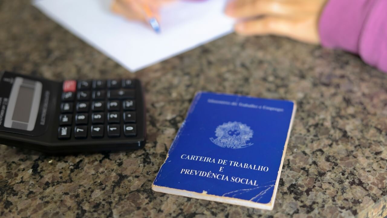 fotografia de uma carteira de emprego e uma calculadora em uma mesa de mármore com uma pessoa apoiada escrevendo em um papel branco