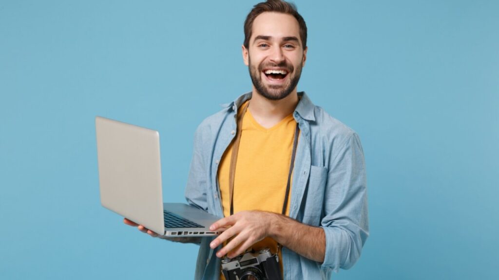 Homem rindo, segurando um laptop, isolado em um fundo azul.