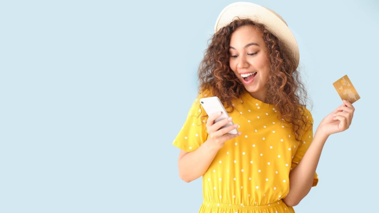 Mulher feliz, segurando um cartão de crédito e um celular em fundo de cor azul clara.