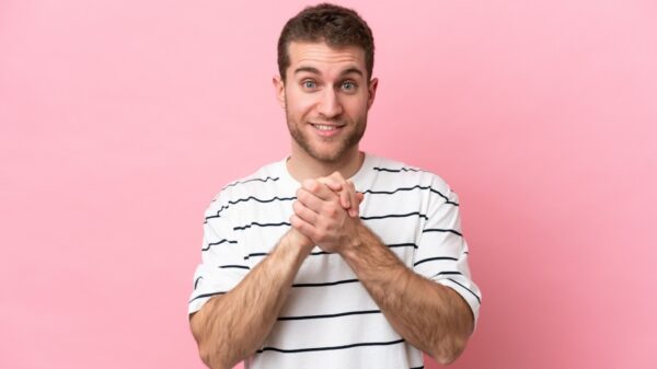 homem com as mãos juntas na altura do peito e sorrindo em um fundo rosa