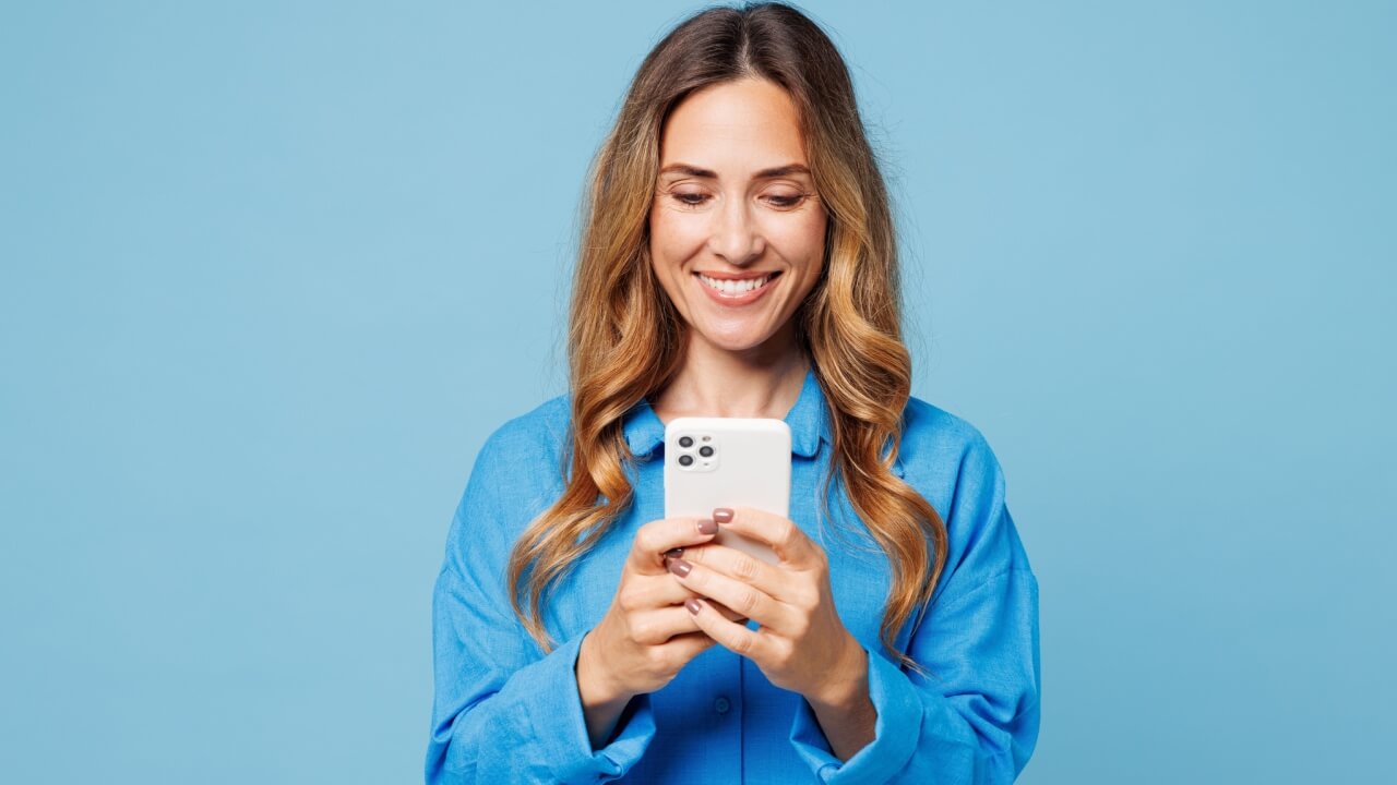 Mulher sorrindo, feliz, alegre, usando roupas casuais, usando um celular, isolada em um estúdio de fundo azul. Conceito de estilo de vida.