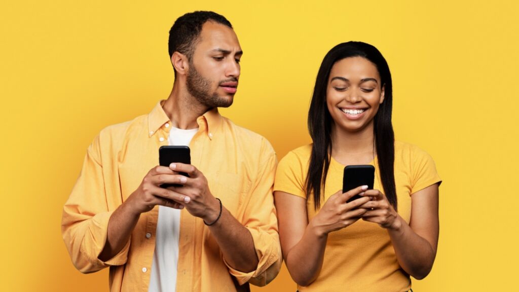 homem com um celular na mão e olhando por cima do ombro da mulher para enxergar o celular delas. ambos de amarelo em um fundo amarelo