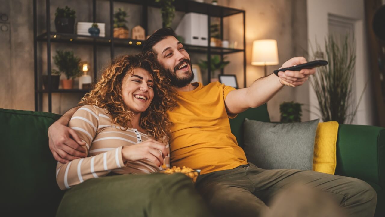 casal heterossexual sentado no sofá com o homem segurando o controle remoto da tv