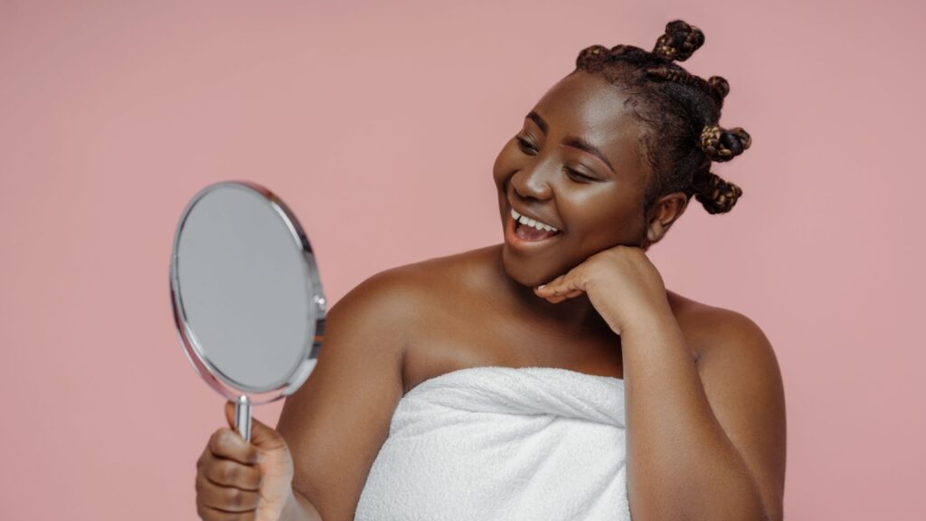 mulher negra sorrindo segurando um espelho em fundo rosa