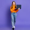 menina vestida com roupa laranja segurando um notebook em fundo roxo