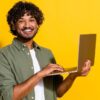 Retrato fotográfico de um homem de cabelos cacheados segurando um computador, isolado em um fundo amarelo.